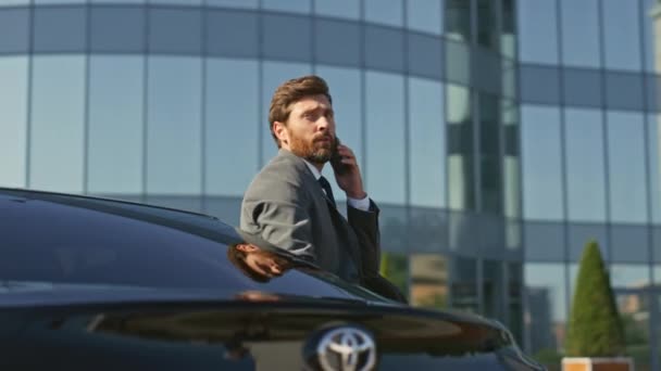 Successful entrepreneur getting out car premium class talking cell phone close up. Calling bearded businessman opening door expensive automobile arrived to modern office building. Camera moving around - Footage, Video