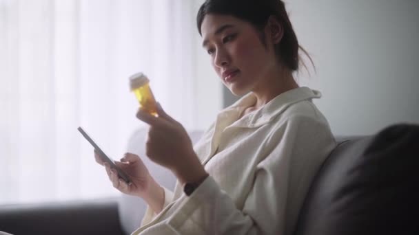 Больная азиатка, держащая в руках бутылку с таблетками, читает инструкции или рецепт на лекарства при гриппе на диване в гостиной дома, здравоохранение и медицина концепции - Кадры, видео