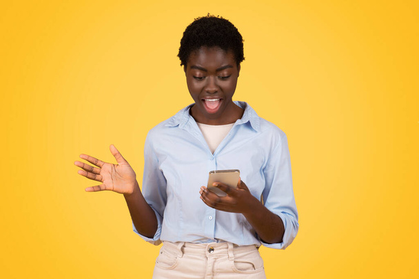 Glad shocked teen black lady student, typing on smartphone, in social networks, enjoy win, isolated on yellow background, studio. Surprise, read message, good news and app - Fotó, kép