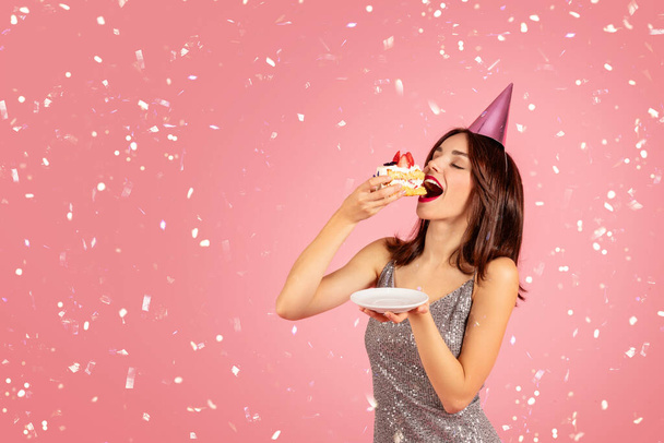 Cheerful pretty millennial caucasian brunette woman in party cap, dress eat cake, isolated on pink background, studio. Celebration birthday party with confetti, fun and fashion - Foto, Imagem