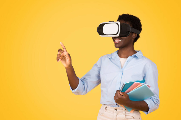 A black woman student, engaged in futuristic learning with VR glasses with books, expression one of wonder and curiosity, set against yellow background symbolizing innovation in education - Fotografie, Obrázek