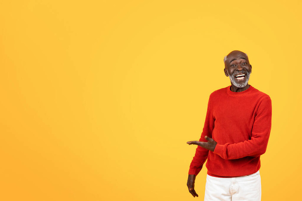 Joyful senior Black man in a red sweater laughing and presenting something to his side, standing against a bright yellow studio background with copy space, ad, offer and sale - Photo, image