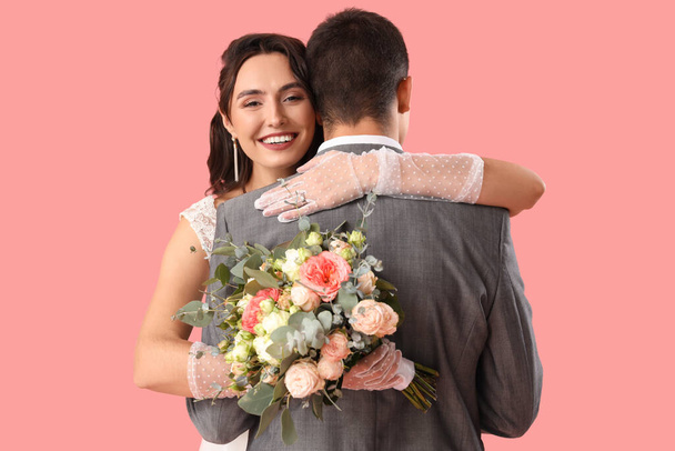 Young wedding couple with bouquet of flowers hugging on pink background - Foto, afbeelding