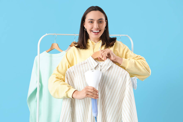 Pretty young woman steaming clothes with modern steamer on blue background - Valokuva, kuva