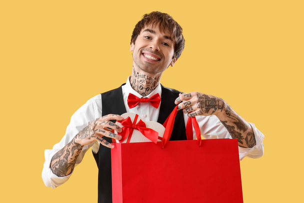 Handsome young man putting gift for Valentine's day in shopping bag on yellow background - Photo, Image