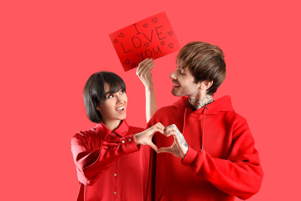 Young couple with gift card for Valentine's day and making heart with hands on red background - 写真・画像
