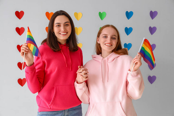 Young lesbian couple with LGBT flags against rainbow hearts on light background. Valentine's Day celebration - Photo, image