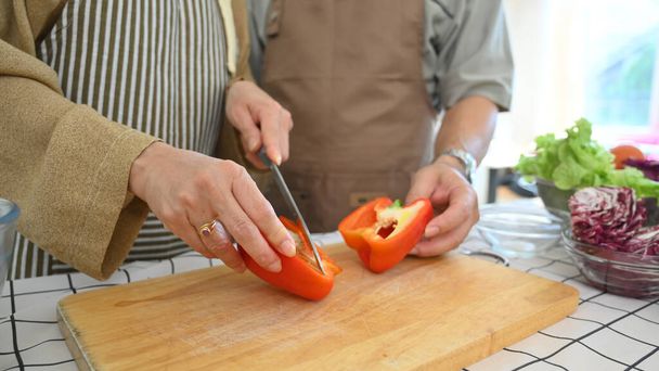 Вырезанный выстрел пожилая женщина резки колокольчик перец на деревянной доске при подготовке здорового салата на кухне - Фото, изображение