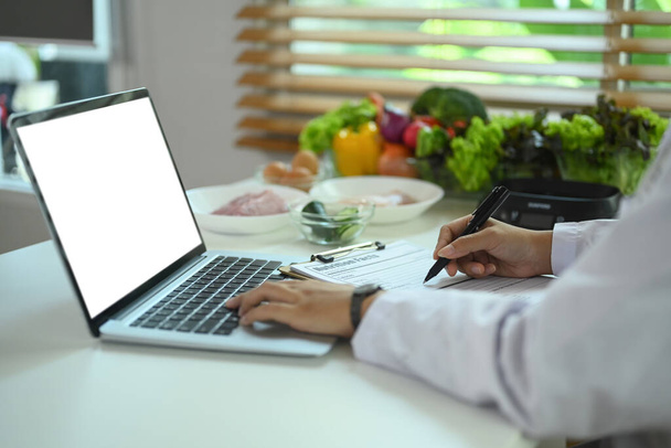 Zobacz zbliżenie profesjonalny dietetyk za pomocą laptopa i przepisu na biurko - Zdjęcie, obraz