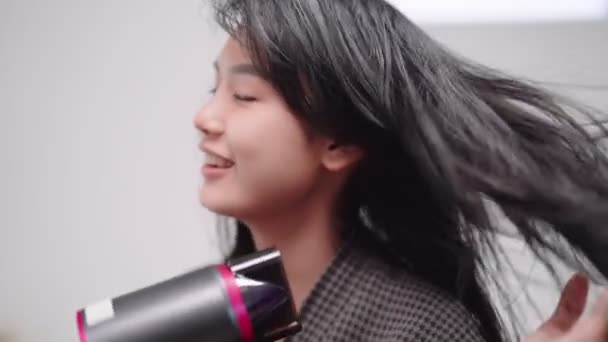 An Asian woman blow-drying her hair in front of a large mirror after washing and styling it at home. Capture the essence of beauty and self-care. High quality 4k footage - Materiał filmowy, wideo