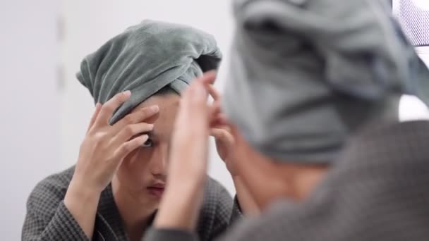 Asian woman is examining her face for blemishes and acne in front of a bathroom mirror, preparing for a shower. Capture skincare routine and self-care moments for a radiant image - Filmagem, Vídeo