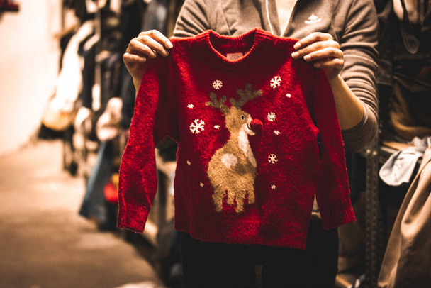 Warmer rotgestrickter Weihnachtspullover mit einem Bild eines niedlichen Hirsches in Frauenhand. Die Wahl der Kleidung für ein Kind, Kind. Junge Mutter kauft Kleidung für ein Neujahrsgeschenk. Winterschlussverkauf. Vintage-Geschäft. - Foto, Bild