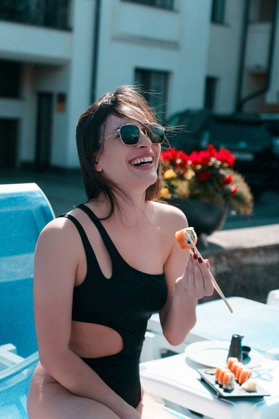 A young woman in a black swimsuit eats a Philadelphia roll near the pool. Concept of rolls and sushi. Relaxation near the pool. Summer season - Foto, Bild
