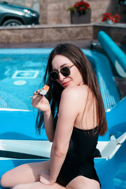 A young woman in a black swimsuit eats a Philadelphia roll near the pool. Concept of rolls and sushi. Relaxation near the pool. Summer season - Fotó, kép