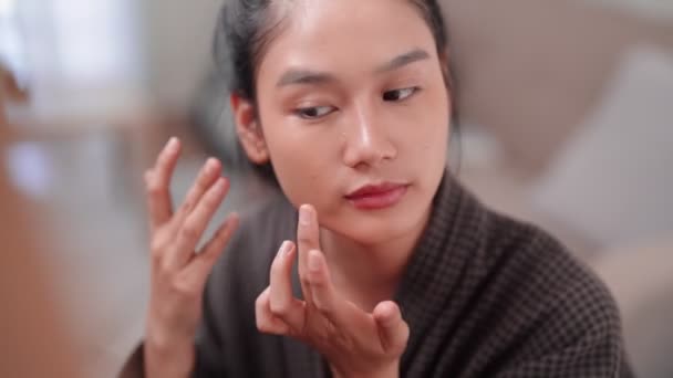 Asian woman applying concealer on her face in front of a large mirror after washing and styling her hair at home. Capture her beauty routine. High quality 4k footage - Filmati, video