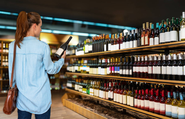 Rear view of woman customer stands in front of shelves with wine bottles choosing wine in liquor store. - Fotoğraf, Görsel