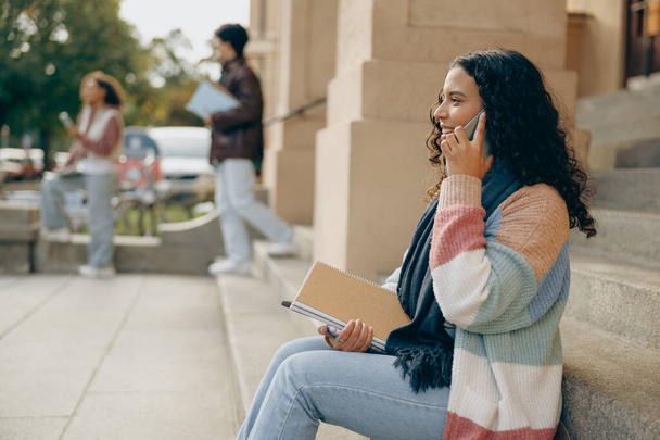 Улыбающаяся студентка разговаривает по телефону сидя на лестнице рядом с университетом на фоне друзей - Фото, изображение