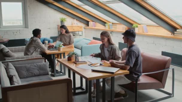 Full shot of young Caucasian male and female freelancers in casual outfits sitting in coworking with laptop and tablet and discussing document draft, black man and Asian woman talking in background - Materiaali, video