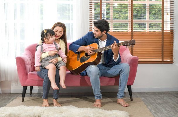 young interracial parent with a little daughter sitting on sofa playing guitar,singing a song,family spend leisure time together in the living room,concept of family,relationship,the love of a family - Photo, image