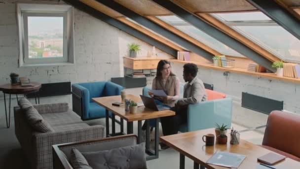 Full zoom shot of young Asian woman and African American man sitting together on couch in loft style coworking office, laptop on table, holding presentation and discussing financial data - Кадры, видео