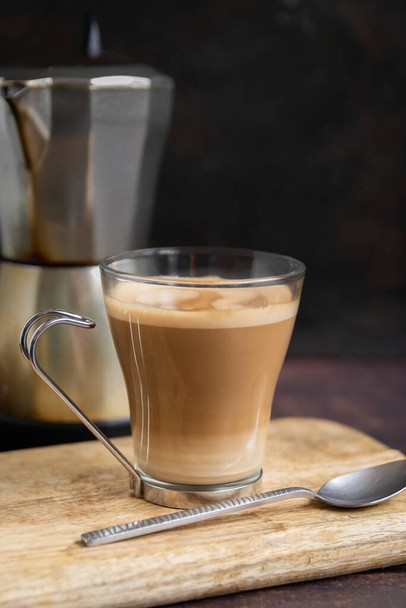 Filiżanka kawy z mlekiem i łyżeczką na drewnianym stole i włoski dzbanek do kawy w tle. Pionowe - Zdjęcie, obraz