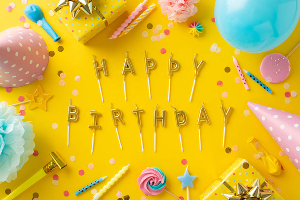 Schattig feest. Overhead shot van vrolijke setup, lolly 's, geschenken, party caps, Happy Birthday brief kaarsen, ballon, pijp, confetti, bloemen op levendige gele achtergrond - Foto, afbeelding