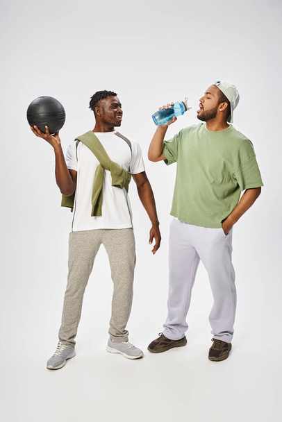 happy african american man holding ball and looking at friend drinking water on grey background - Foto, Bild