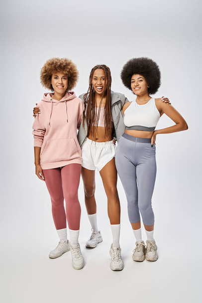 vreugdevolle Afrikaanse amerikaanse vrouwen in sportkleding staan samen op een grijze achtergrond, Juneteenth concept - Foto, afbeelding