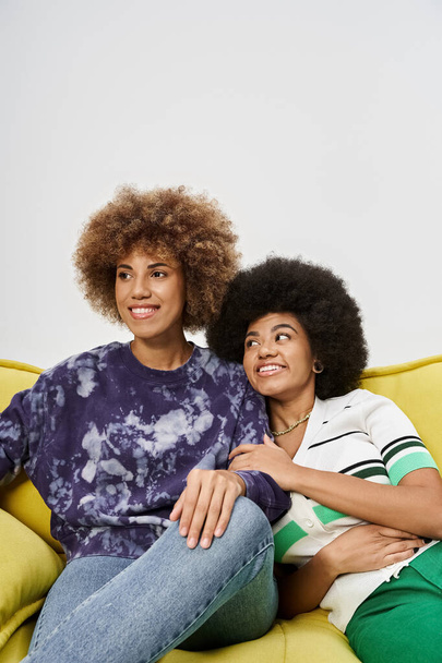 positive and curly african american female friends sitting on yellow sofa on grey background - 写真・画像