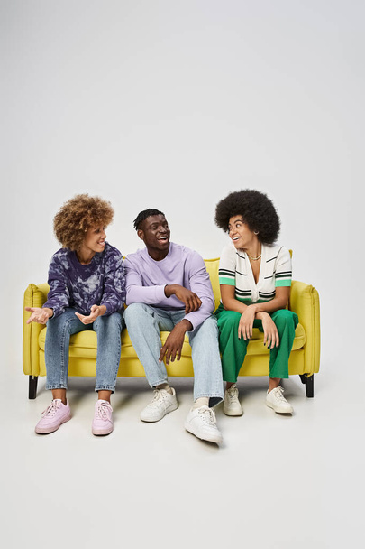 joyful and curly african american friends chatting and sitting on yellow sofa on grey background - Fotó, kép