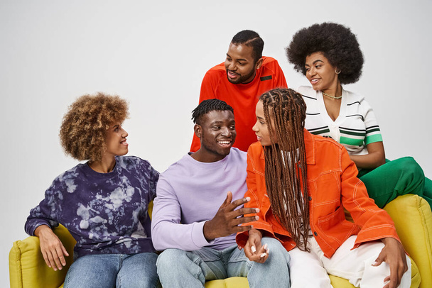 smiling african american friends in colorful casual attire chatting and sitting on yellow couch - Photo, Image