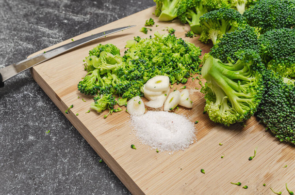 Green broccoli vegetable with garlic and salt on wooden board. Sharp kitchen knife. Dark background - Photo, Image