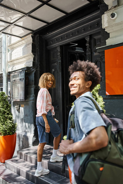 счастливый африканский американец мужчина рядом с подругой входит молодежный хостел на улице города, развлечения и путешествия - Фото, изображение