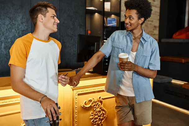 alegre afroamericano hombre con taza de papel hablando con un amigo sonriente en recepción en el albergue - Foto, Imagen