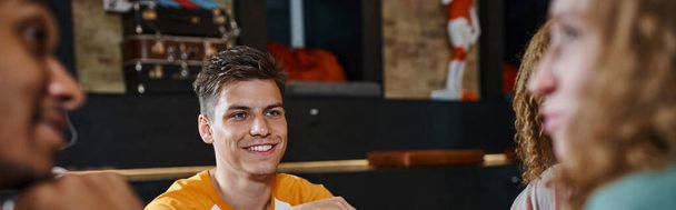 młody stylowy człowiek uśmiechnięty w pobliżu wielokulturowych kumpli rozmawiających w holu hostelu studenckiego, baner - Zdjęcie, obraz