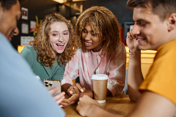 opgewonden vrouw tonen smartphone naar gelukkig multi-etnische vrienden in lounge cafe van studenten hostel - Foto, afbeelding