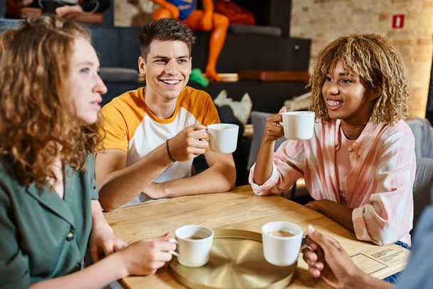 веселые мультикультурные друзья, держащие чашки кофе и беседующие в общежитии кафе, путешествия и дружбы - Фото, изображение