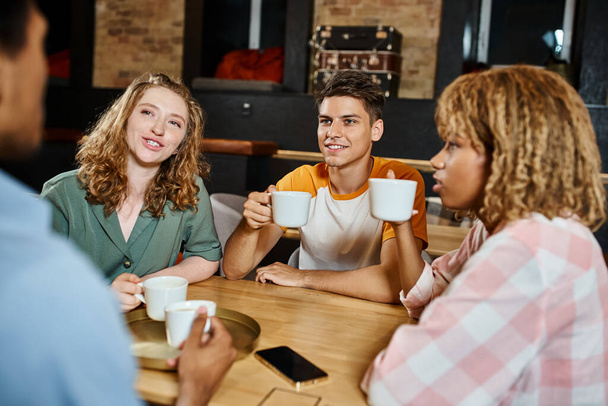 positive multiethnic friends with coffee cups talking in lounge cafe of students hostel, travelers - Photo, Image