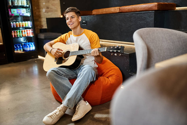 wesoły młody człowiek siedzi na krześle worek fasoli i gra na gitarze w lobby nowoczesnego hostelu studenckiego - Zdjęcie, obraz