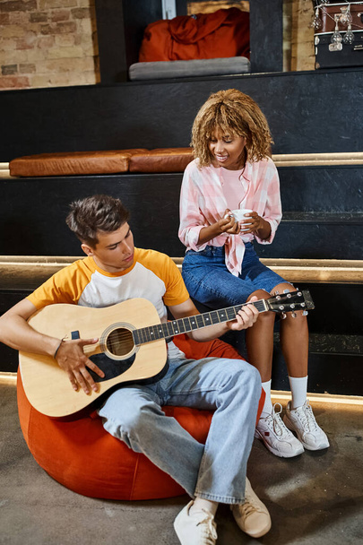 young man playing guitar to pleased african american girlfriend with coffee cup in lounge of hostel - Photo, Image
