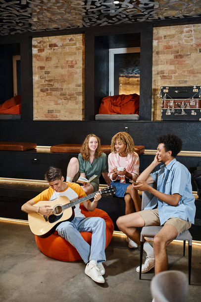 nuori mies soittaa kitaraa iloinen monikansallinen ystäviä lounge opiskelijoiden hostelli, matkustajat - Valokuva, kuva