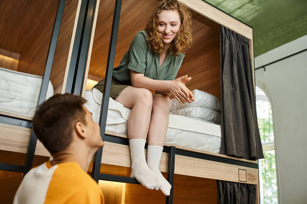 весела молода жінка сидить на двоповерховому ліжку і дивиться на хлопця в затишному гуртожитку - Фото, зображення