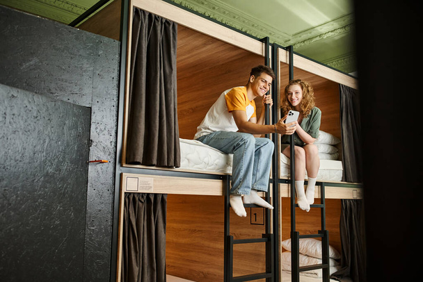 vrolijke jonge vrouw tonen smartphone naar vriendje op dubbeldekker bedden in gezellige hostel kamer - Foto, afbeelding