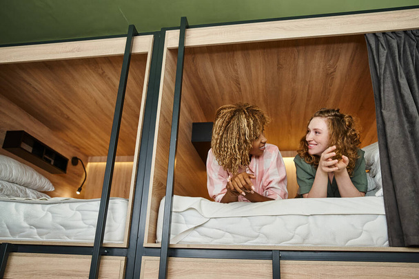 zorgeloze multiculturele vriendinnen liegen en praten op dubbeldekker bed in moderne studentenherberg - Foto, afbeelding