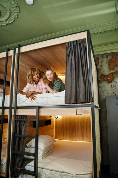 νεανικές ενθουσιασμένες φίλες ξαπλωμένες σε διώροφο κρεβάτι και κοιτώντας μακριά στο σύγχρονο ξενώνα των μαθητών - Φωτογραφία, εικόνα