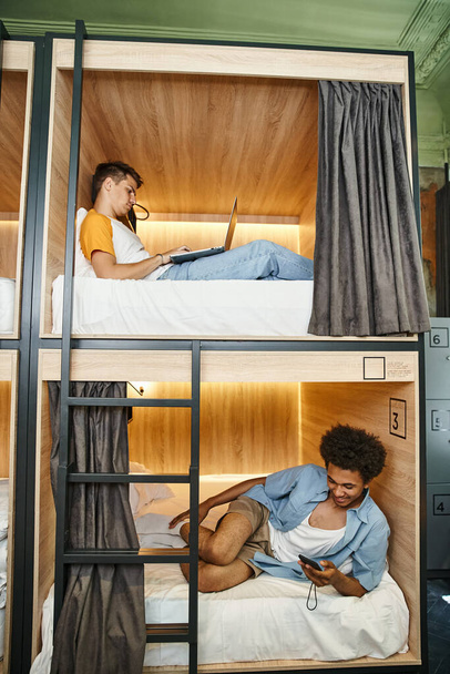 wielokulturowi studenci korzystający z gadżetów na piętrowych łóżkach we współczesnym schronisku młodzieżowym, podróżnicy - Zdjęcie, obraz