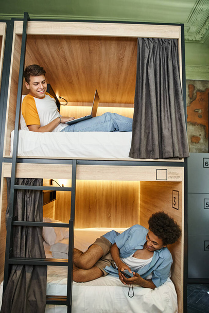 χαρούμενη πολυεθνική συγκάτοικοι που βρίσκονται με φορητό υπολογιστή και smartphone σε διώροφα κρεβάτια σε ξενώνα - Φωτογραφία, εικόνα
