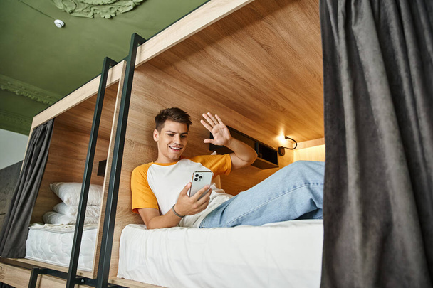 alegre estudiante saludando la mano durante la videollamada en el teléfono inteligente en la cama de dos pisos en el albergue - Foto, imagen