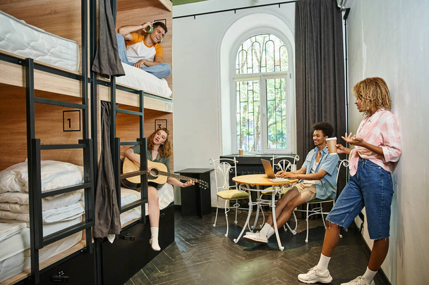 ocio de alegres amigos multiétnicos descansando en acogedora habitación de albergue juvenil, estudiantes viajeros - Foto, Imagen