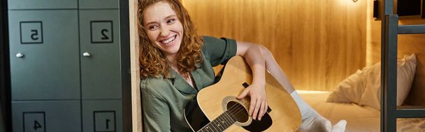 radostná mladá žena hrající na akustickou kytaru na double-decker posteli v pohodlném hostelu pokoj, banner - Fotografie, Obrázek